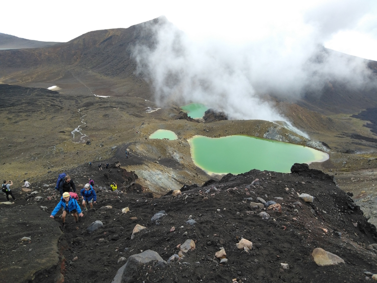 Photo shows children in blue jackets climbing in volcanic areas with green-blue lakes behind them and steam rising from the ground.